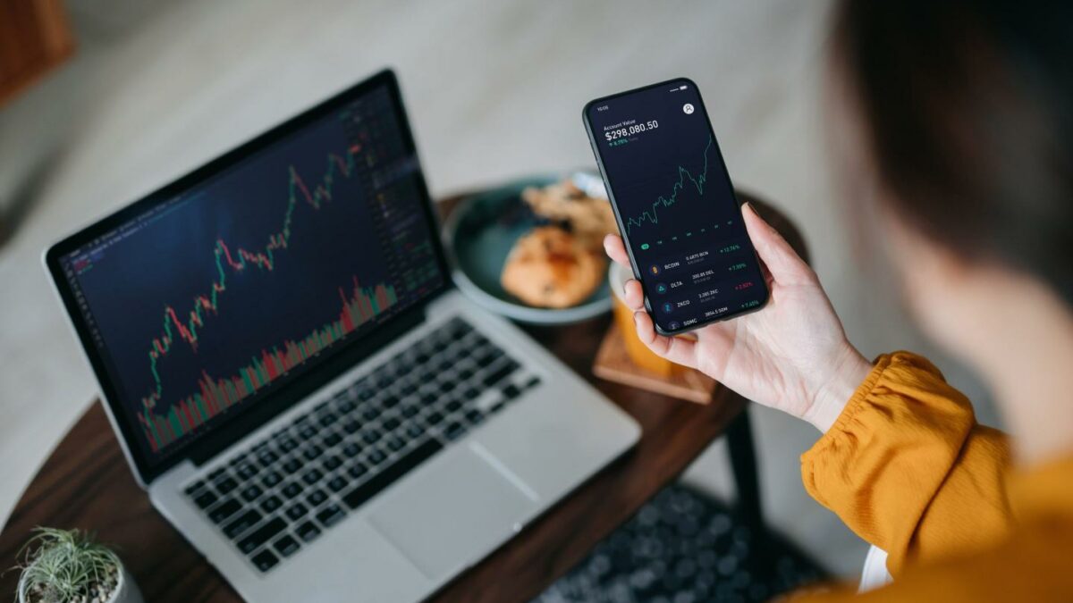 Woman holds phone displaying crypto price chart with a laptop in front of her also showing crypto price chart.