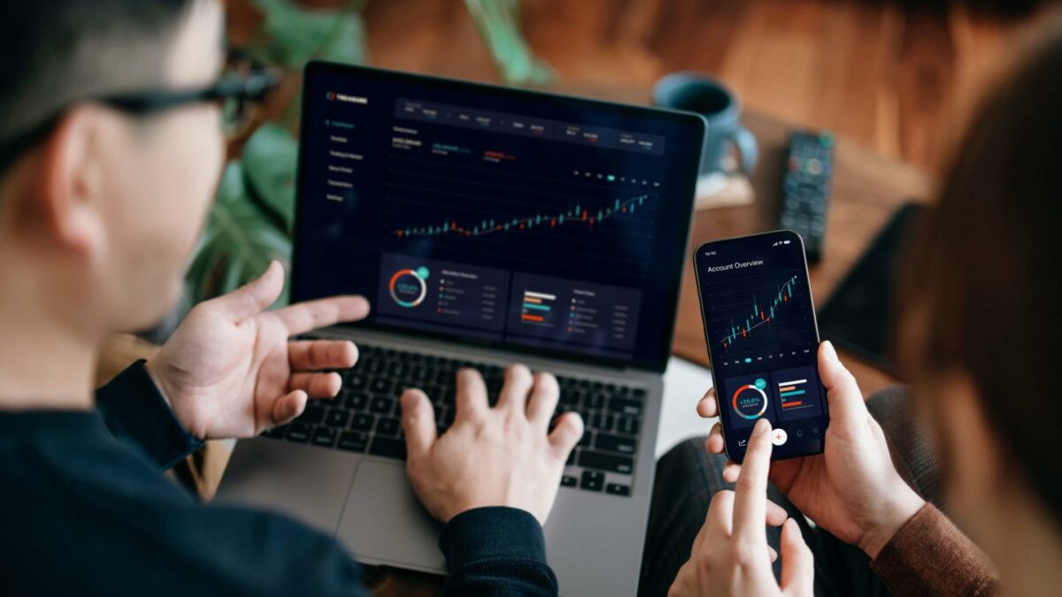 A man on the left evaluates trading charts on his laptop while a woman on his right does the same thing on her phone.