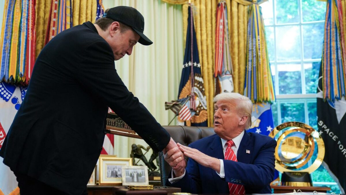 Trump, sitting at his desk in the Oval Office, shakes Elon Musk's hand.