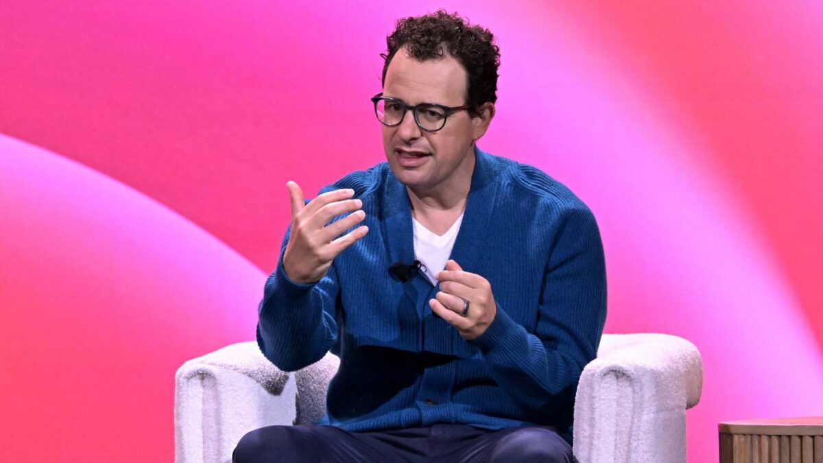 Dario Amodei sits in a white chair in front of a pink background and speaks animatedly.