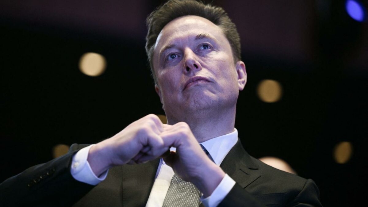 Elon Musk sits with his fists together, looking up.