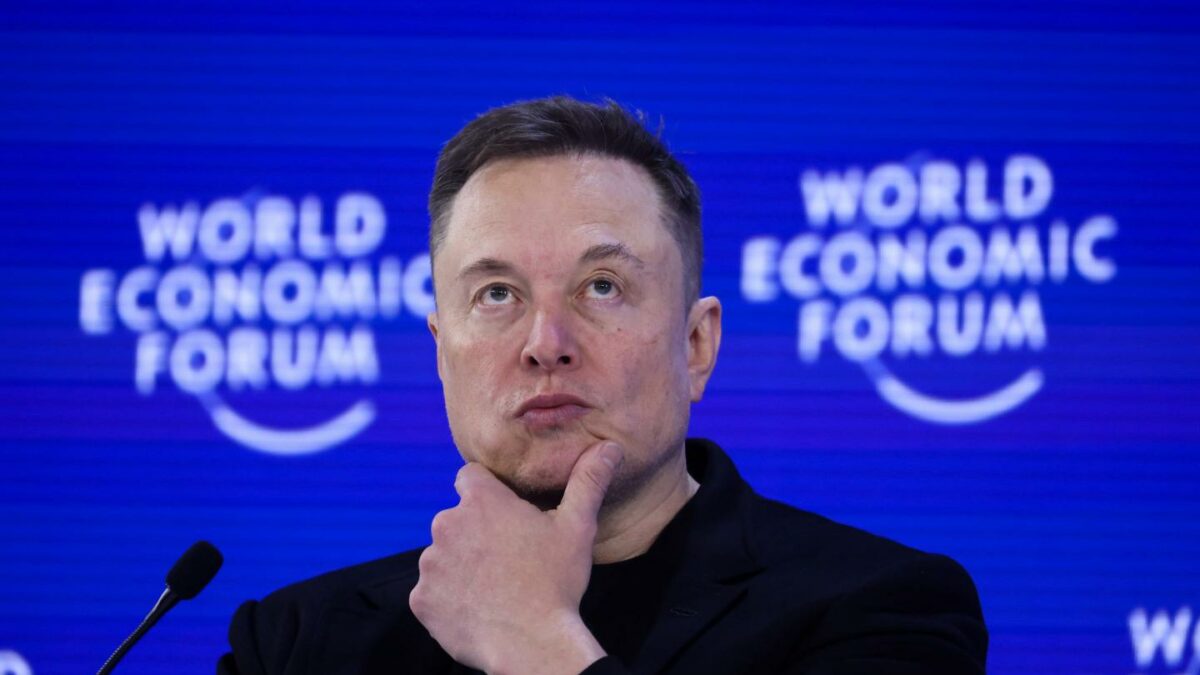 Elon Musk puts one hand to his chin and he looks up. He is in front of a blue "World Economic Forum" background.