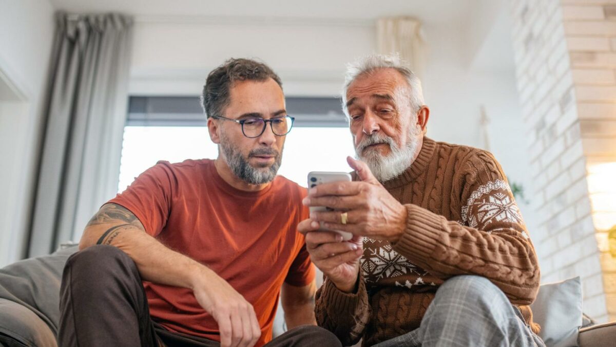 A younger man and an older man sit next to each other on a couch, looking down at a cell phone.