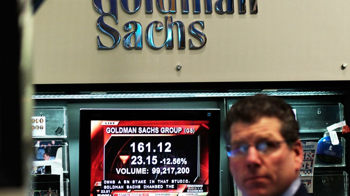 A television screen showing Goldman Sachs' stock price plays in front of a Goldman Sachs sign